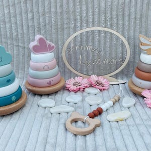 May include: Three wooden stacking toys with different color schemes and designs. The first toy is blue and white with a cloud shape on top. The second toy is pink and white with a heart shape on top. The third toy is brown and white with a butterfly shape on top. A wooden teething ring and a sign that says "from Lyzamora" are also in the image.