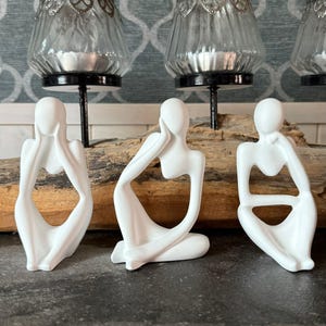 May include: Three white abstract human figure sculptures in different poses. The figures are on a piece of wood, with glass candle holders with silver accents behind them. The sculptures have a minimalist, modern design.