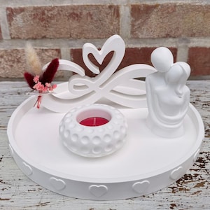 May include: A white decorative tray with a heart-shaped design, featuring a candle holder, a sculpture of embracing figures, and a floral arrangement. The tray has a heart-shaped border and a red candle.