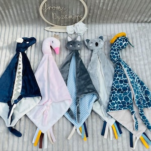 Puede incluir: Cinco juguetes de peluche para bebés con diferentes diseños de animales. Los juguetes son azules, rosas, grises, azules claros y verde azulado. Cada juguete tiene una manta suave y blanca unida a él. Los juguetes están dispuestos en una fila sobre una superficie gris.