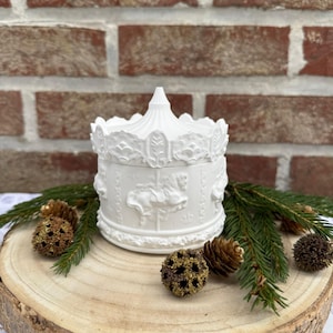 Op de afbeelding: Een witte keramische sieradendoos met carrouselthema en een gedetailleerd ontwerp. De doos heeft carrouselpaarden en decoratieve versieringen. Het deksel heeft een conische bovenkant. De doos wordt gepresenteerd op een houten schijf met dennennaalden en decoratieve dennenappels.