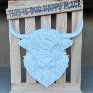 May include: A white Highland cow head sculpture mounted on a wooden frame. The sculpture has detailed fur and horns. The text "THIS IS OUR HAPPY PLACE" is above the cow head.