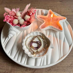 Peut inclure: Un plateau blanc en forme de coquillage avec un bol en forme de coquillage rempli de fleurs séchées roses, une bougie en forme d'étoile de mer et un plat en forme de coquillage contenant des bracelets de perles. La bougie est allumée.
