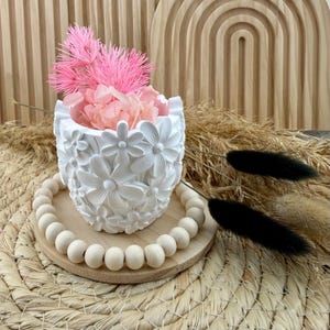 May include: A white cylindrical planter featuring a raised floral relief pattern, containing pink dried flowers. The planter rests on a circular wooden base surrounded by a ring of light-colored wooden beads, accompanied by two black dried stems.