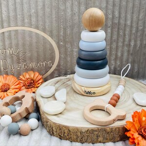 May include: A wooden stacking toy with gray and white rings, a wooden teether with a bear face, and a wooden bead necklace with gray, white, and brown beads. The toy is on a wooden tray with the word "Tryco" printed on it.