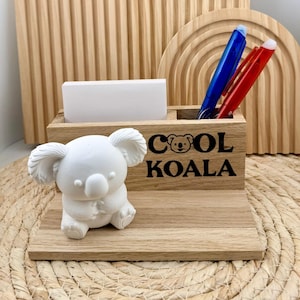 Könnte beinhalten: Ein Schreibtisch-Organizer aus Holz mit einer weißen Koala-Figur, einem Notizblock und Stifthaltern. Auf der Vorderseite des Organizers steht der Text "COOL KOALA". In den Stifthaltern befinden sich zwei Stifte, einer blau und einer rot.