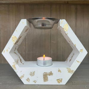 Può includere: Un portacandele geometrico bianco con accenti in foglia d'oro. Ha una forma esagonale con una ciotola di metallo nella parte superiore e una candela tealight accesa nella parte inferiore. Il diffusore per aromaterapia è progettato per oli essenziali.