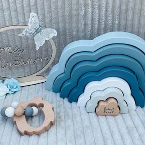 May include: A wooden rainbow toy with blue and white painted clouds. The rainbow is made of five stacked arches. There is a small wooden heart with the text "label label" on it. There is also a wooden sign with the text "from Dyzamora" and a butterfly.