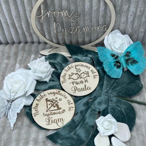 May include: Two wooden plaques with engraved text and images. The first plaque is brown and has the text 'Notre bebe poissons est la' and 'Pisces' with a fish symbol and the name 'Paula'. The second plaque is brown and has the text 'Notre bebe sagittaire est la' and 'Sagittarius' with an archer symbol and the name 'Liam'.