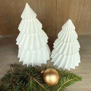 Könnte beinhalten: Zwei weiße Keramik-Weihnachtsbäume mit einem geschichteten Design. Eine goldene Kugel und Tannenzweige befinden sich im Vordergrund. Die Bäume haben ein strukturiertes, plissiertes Aussehen, das auf eine papierartige Konstruktion hindeutet. Die Szene erinnert an ein festliches Urlaubsthema.