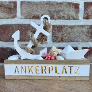 Könnte beinhalten: Weiße Holzdekoration mit maritimem Thema, mit Anker, Seil, Muscheln und Papierboot. Die Basis hat den Schriftzug "ANKERPLATZ" ausgeschnitten, mit einem Hintergrund aus Ziegeln.