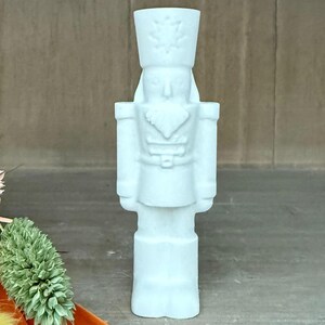 May include: A light blue nutcracker ornament. The figurine is in the shape of a soldier with a tall hat and a detailed uniform. The nutcracker is standing upright and has a detailed face. The background is a neutral brown.