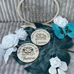 May include: Two wooden plaques with engraved text and images. The first plaque reads 'Notre bébé gémeaux est là Gemini II Emma.' The second plaque reads 'Notre bébé cancer est là Cancer Theresa.' Both plaques have a cartoon image of a baby with a zodiac symbol. The plaques are on a green velvet surface with white roses and a blue butterfly.