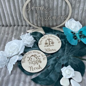 May include: Two wooden round plaques with engraved text and zodiac symbols. The first plaque says 'Notre bébé vierge est là' with a Virgo symbol and the name 'Noah'. The second plaque says 'Notre bébé balance est là' with a Libra symbol and the name 'Klara'. Both plaques are on a teal green surface with white roses and a silver butterfly.