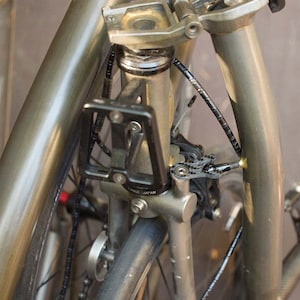 May include: Close-up of a bicycle frame with a black water bottle cage and various components. The frame is a metallic gray color, and the image highlights the bike's structure and details. The text "TANGE JAPAN" is visible on the frame.