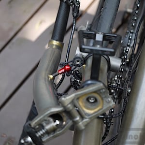 May include: Close-up of a bicycle frame with a red component and black cables. The frame is metallic gray, with a curved tube and various mechanical parts visible. The chain and tire are also in view.