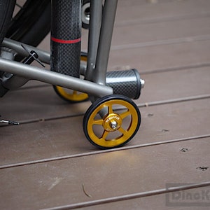 May include: A close-up of a folding bicycle with gold-coloured wheels. The wheels have a five-spoke design and black rubber tyres. The frame is grey, and a carbon fibre component is visible. The bicycle is on a wooden surface.