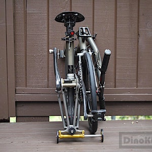 May include: A folded olive green folding bicycle with black tyres and seat. The bicycle has a black handlebar and a gold-coloured wheel at the bottom. The bike is positioned on a wooden deck against a brown wooden fence.