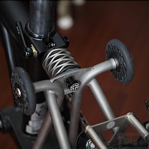 May include: Detail of a bicycle's rear suspension. It shows a silver spring, black and silver parts, and small black wheels. The design suggests a folding bike, emphasizing compact size and functionality, with measurements in cm.
