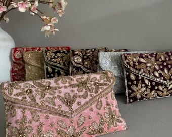Puede incluir: Una colección de bolsos de mano de terciopelo en varios colores, incluyendo rosa, dorado, negro, plateado y burdeos. Cada bolso está adornado con intrincados abalorios y bordados dorados, con diseños florales y de cachemira. Los bolsos se exhiben sobre una superficie neutra.