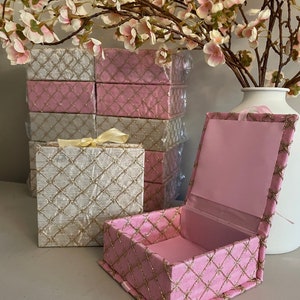 May include: Pink and white decorative boxes with gold lattice embroidery. One box is open, revealing a pink interior. A stack of similar boxes is in the background, along with a vase of pink flowers. The boxes are tied with a yellow ribbon.