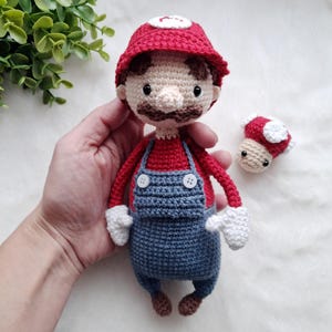 Peut inclure: Poupée Mario au crochet, portant un chapeau rouge avec un cercle blanc, une chemise rouge, une salopette bleue et des chaussures marron. Un petit champignon au crochet avec un chapeau rouge et des taches blanches est également présent. La poupée mesure environ 15 cm de haut.