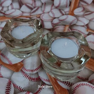 Puede incluir: Dos portavelas de vidrio transparente con bordes ondulados, cada uno con una vela de té blanca. Los portavelas están sobre una superficie cubierta de pelotas de béisbol.