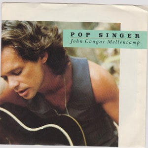 May include: A black and white photo of a man with long brown hair, wearing a black tank top and a silver necklace. He is holding a black acoustic guitar and looking down at it. The text "POP SINGER John Cougar Mellencamp" is printed in white on a light blue background at the top of the image.