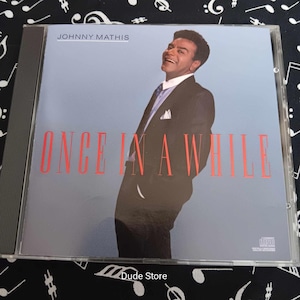 May include: A CD case featuring Johnny Mathis. The album cover has a light blue background with the text "ONCE IN A WHILE" in red. Johnny Mathis is pictured smiling, wearing a black suit and tie. The CD case is in a clear plastic case.