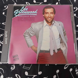 Puede incluir: Una caja de CD con el álbum "Greatest Hits" de Lee Greenwood. La portada tiene un fondo rosa con el nombre del artista en turquesa y blanco. La imagen muestra a un hombre con una blazer gris, camisa blanca y pantalones rojos.