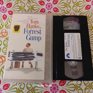 May include: A VHS tape of the movie Forrest Gump starring Tom Hanks. The front of the case features a photo of Tom Hanks as Forrest Gump sitting on a bench. The text on the case reads "The world will never be the same once you've seen it through the eyes of Forrest Gump. Tom Hanks is Forrest Gump."