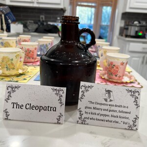 May include: A dark brown glass jug with a handle sits on a table with floral teacups and saucers. Two cards read "The Cleopatra" and a quote about Cleopatra. The teacups have yellow, pink, and blue floral designs.