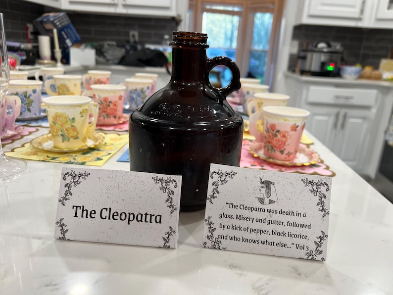 May include: A dark brown glass jug with a handle sits on a table with floral teacups and saucers. Two cards read "The Cleopatra" and a quote about Cleopatra. The teacups have yellow, pink, and blue floral designs.