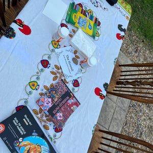 May include: A collection of art supplies on a white tablecloth with a colorful floral design. The supplies include paint markers, crayons, marbled paper, glue, and blank cards, suggesting a crafting or art project.