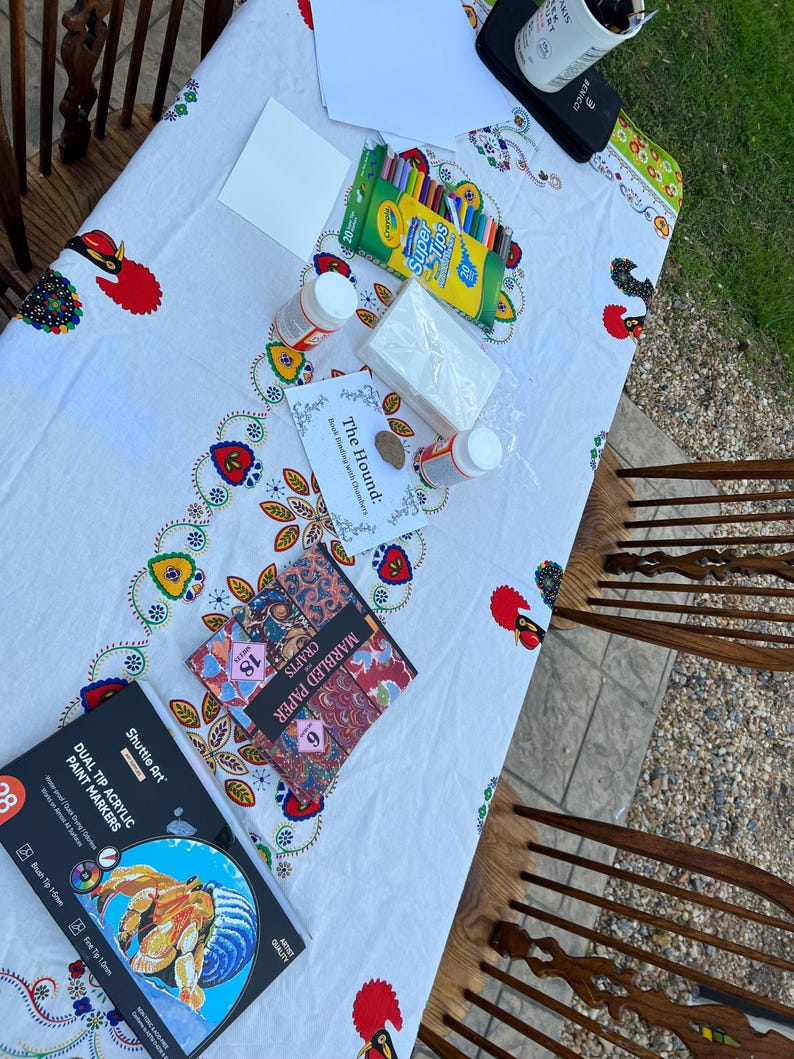 May include: A collection of art supplies on a white tablecloth with a colorful floral design. The supplies include paint markers, crayons, marbled paper, glue, and blank cards, suggesting a crafting or art project.