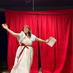 May include: A person in a white dress with a red sash and blue accents, holding a paper, stands in front of a red curtain. The person is smiling and has their arm raised.