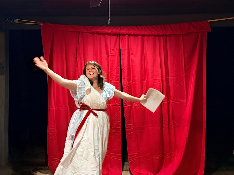 May include: A person in a white dress with a red sash and blue accents, holding a paper, stands in front of a red curtain. The person is smiling and has their arm raised.