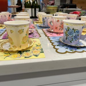 May include: A collection of floral-decorated teacups and saucers arranged on a white surface. The cups feature various floral designs in shades of yellow, pink, and blue. Each cup sits on a matching saucer and decorative placemat, creating a charming tea party setting.