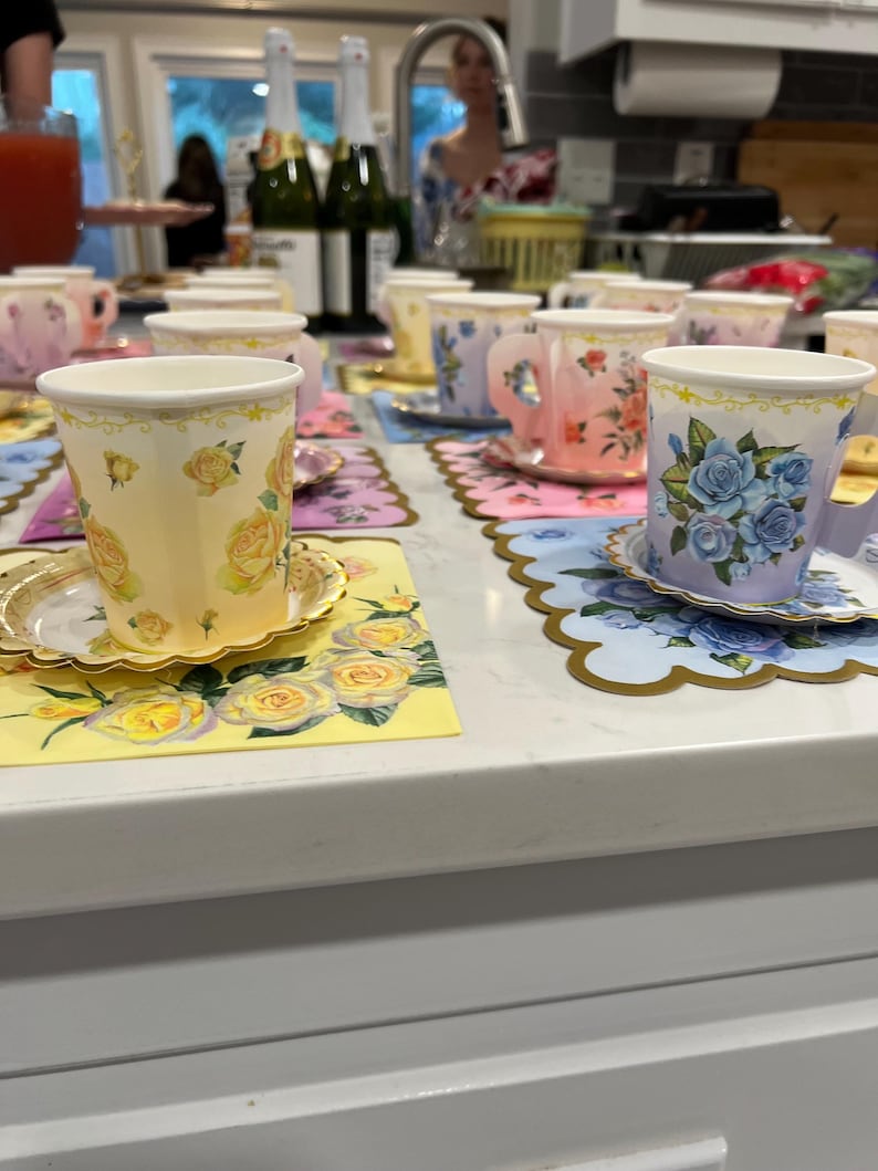 May include: A collection of floral-decorated teacups and saucers arranged on a white surface. The cups feature various floral designs in shades of yellow, pink, and blue. Each cup sits on a matching saucer and decorative placemat, creating a charming tea party setting.