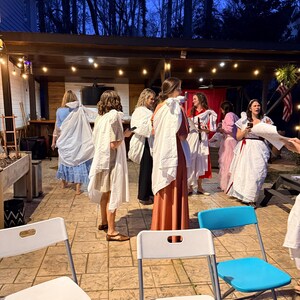 May include: A group of people in white and colorful robes are gathered outdoors under a wooden pergola. The scene is lit by string lights and the sky is dark. White and blue folding chairs are in the foreground.