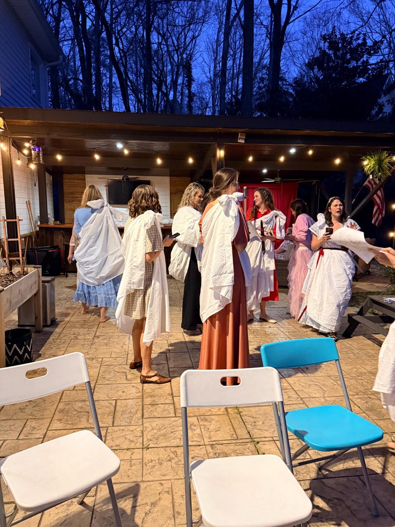 May include: A group of people in white and colorful robes are gathered outdoors under a wooden pergola. The scene is lit by string lights and the sky is dark. White and blue folding chairs are in the foreground.