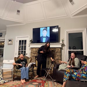 May include: A group of people are gathered in a living room, with a television displaying a person in a tuxedo. The room features a patterned rug, a stone fireplace, and various seating arrangements. The people are smiling and laughing.
