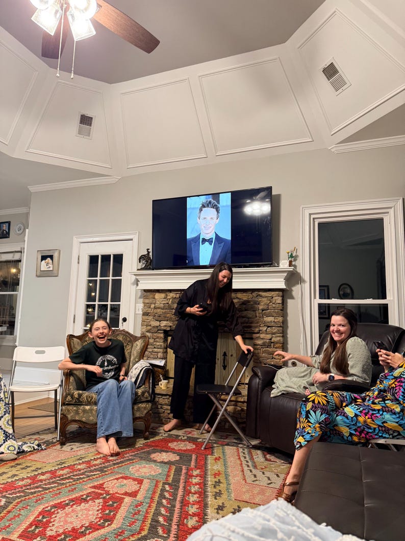 May include: A group of people are gathered in a living room, with a television displaying a person in a tuxedo. The room features a patterned rug, a stone fireplace, and various seating arrangements. The people are smiling and laughing.