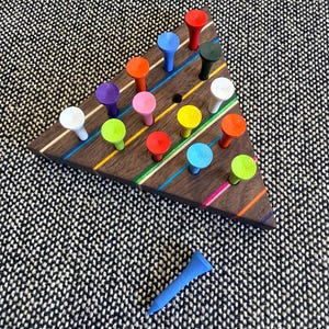 Op de afbeelding: Een driehoekig houten spelbord van donkerbruin hout met diagonale, kleurrijke strepen. Het bord bevat meerdere gaten gevuld met kleine, felgekleurde plastic golftees in de kleuren rood, blauw, oranje, groen, geel, wit en paars.