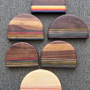 May include: Six semi-circular wooden objects arranged in a cluster. Five pieces are made of dark brown wood, and one is made of light beige wood. Each piece features horizontal, multi-colored thin stripes layered along the flat bottom edge.