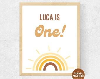 May include: A printable sign with a rainbow and sun design. The text on the sign reads "Luca is One!" in brown and orange lettering.