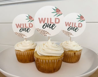 以下が含まれることがあります： 白いフロスティングをかけたカップケーキが3つ。それぞれに「Wild One」とピンクと黒の文字で書かれた丸い紙の円が乗っています。円には花柄とヒョウ柄のパターンがあります。