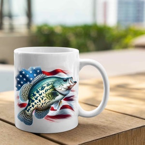 Op de afbeelding: Witte keramische koffiemok met een afbeelding van een baars die voor een Amerikaanse vlag zwemt.