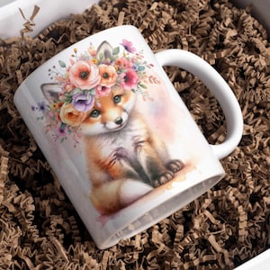 Puede incluir: Una taza de cer&aacute;mica blanca con una ilustraci&oacute;n de acuarela de un zorro con una corona de flores. El zorro est&aacute; sentado sobre un fondo rosa claro con un efecto de acuarela.