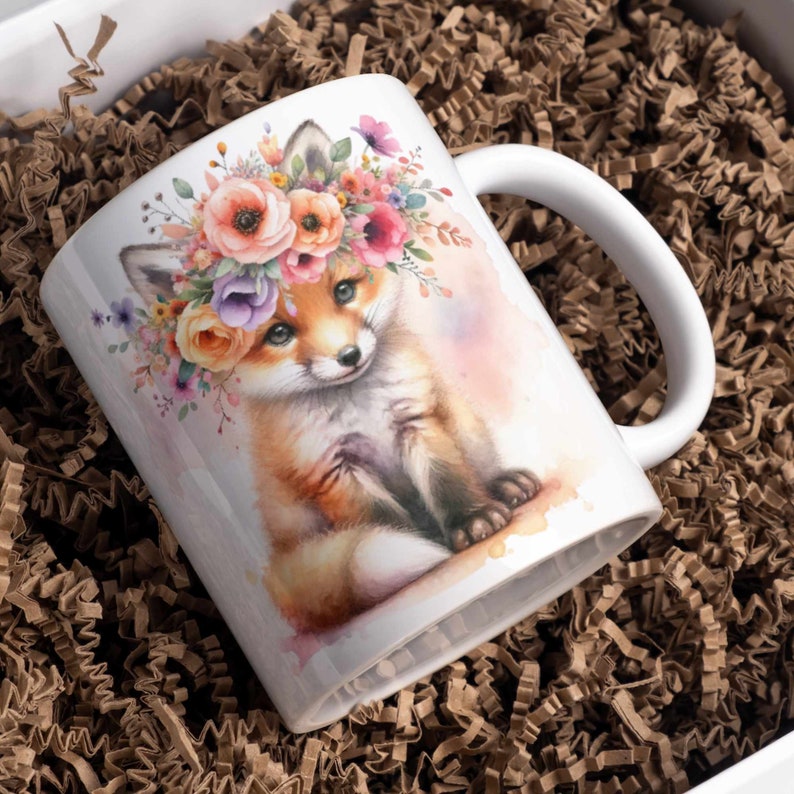 Puede incluir: Una taza de cer&aacute;mica blanca con una ilustraci&oacute;n de acuarela de un zorro con una corona de flores. El zorro est&aacute; sentado sobre un fondo rosa claro con un efecto de acuarela.